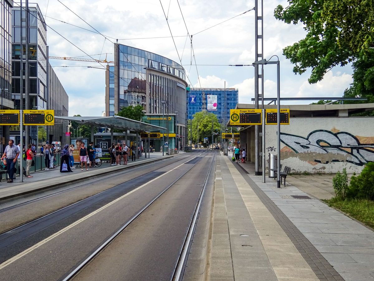 Straßenbahnhaltestelle in einer Innenstadt, auf dem Bahnsteig warten Menschen.