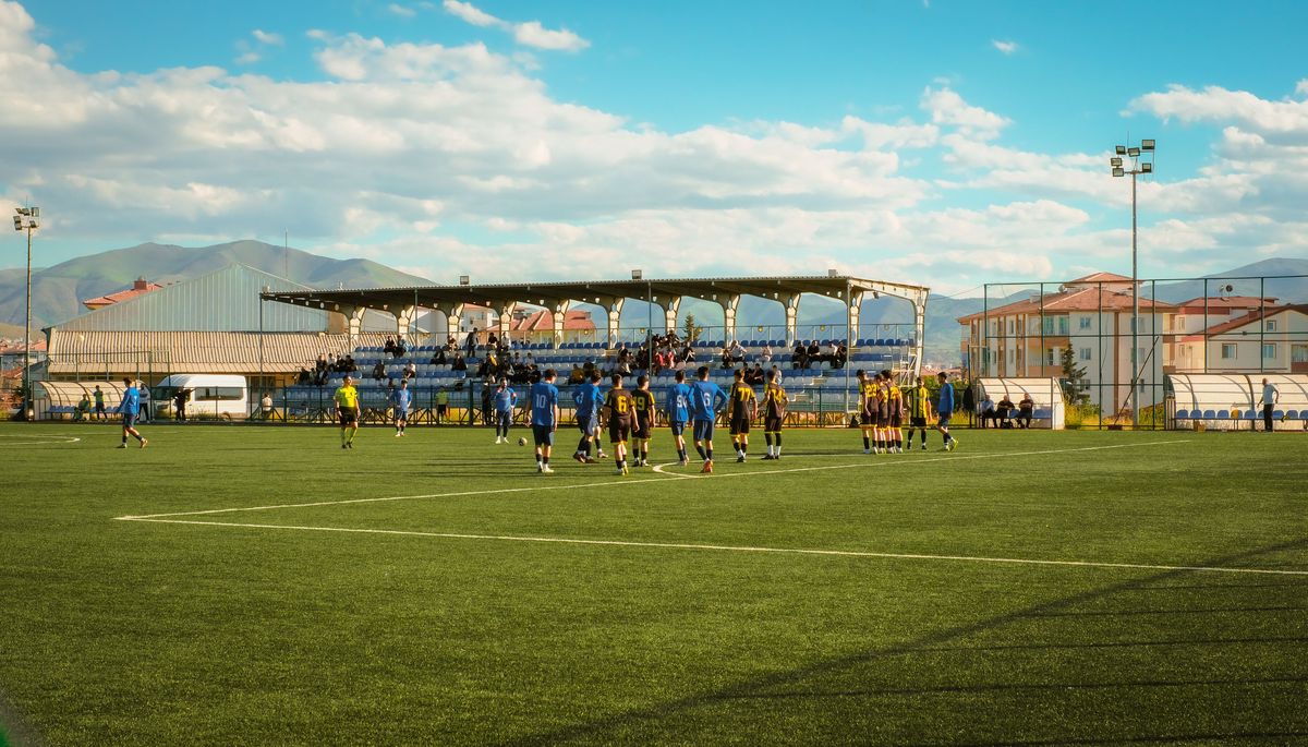 Fußballspiel auf einem Rasenplatz mit kleiner Tribüne, dahinter Berge.